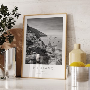 May include: Black and white photograph of Positano, Italy, framed in a light wood frame. The photo shows a coastal town with buildings on a hillside overlooking the sea. The text "Positano, Italy" is printed below the photo.