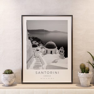 May include: Black and white photograph of Santorini, Greece, featuring whitewashed buildings with blue domed roofs. The image includes the text "SANTORINI" and "GREECE" along with the coordinates "36&deg; 22' N, 25&deg; 26' E".