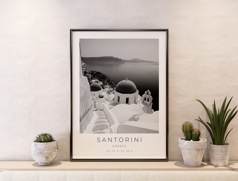 May include: Black and white photograph of Santorini, Greece, featuring whitewashed buildings with blue domed roofs. The image includes the text "SANTORINI" and "GREECE" along with the coordinates "36&deg; 22' N, 25&deg; 26' E".