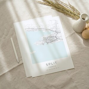 May include: A black and white map print of Split, Croatia, with the city name and coordinates printed at the bottom. The map is on a light beige fabric surface with a white vase and wooden blocks.