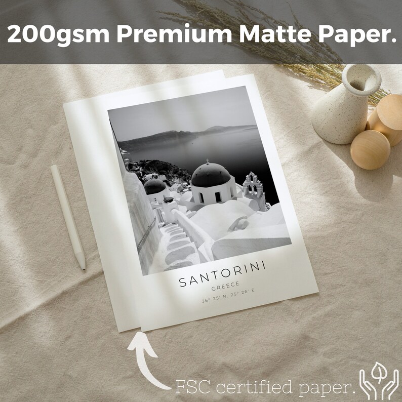 May include: A black and white photograph of Santorini, Greece, printed on 200gsm premium matte paper. The image shows a white-washed building with a dome roof and a staircase leading up to it. The text "SANTORINI" and "GREECE" is printed below the image, along with the coordinates "36 25 N. 25 26 E". The text "FSC certified paper." is printed below the image.