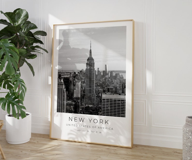 May include: Black and white photograph of the New York City skyline, featuring the Empire State Building. The image includes the text "NEW YORK", "UNITED STATES OF AMERICA", and "40&deg;43' N, 74&deg; 0' W".