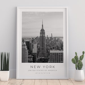 May include: Black and white photograph of the New York City skyline, featuring the Empire State Building. The text "NEW YORK" is printed below the image, along with "UNITED STATES OF AMERICA" and the coordinates "40&deg; 43' N, 74&deg; 0' W".
