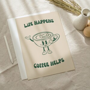 K&ouml;nnte beinhalten: Ein beiger Druck mit einer Cartoon-Kaffeetasse, auf der in dunkelgr&uuml;nen Buchstaben "Life Happens Coffee Helps" steht. Die Kaffeetasse hat Arme, Beine und ein l&auml;chelndes Gesicht. Ein wei&szlig;er Stift, eine kleine Vase und Holzkugeln sind ebenfalls im Bild.