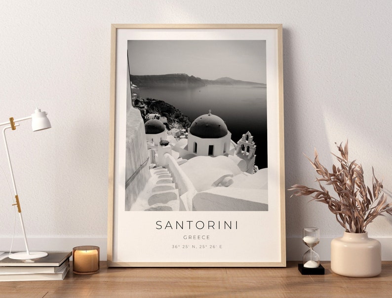 May include: Black and white photograph of Santorini, Greece. The image shows a white building with a dome roof and a staircase leading up to it. The sea and a small island are visible in the background. The text "SANTORINI, GREECE, 36&deg; 25' N, 25&deg; 26' E" is printed below the image.