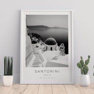 May include: Black and white photograph of Santorini, Greece. The image shows a white building with a domed roof and a staircase leading up to it. The sea and a distant island are visible in the background. The text "SANTORINI" and "GREECE" is printed below the image, along with the coordinates "36&deg; 25' N, 25&deg; 26' E".