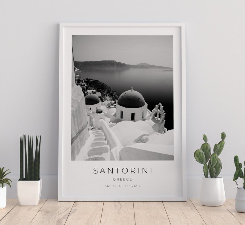 May include: Black and white photograph of Santorini, Greece. The image shows a white building with a domed roof and a staircase leading up to it. The sea and a distant island are visible in the background. The text "SANTORINI" and "GREECE" is printed below the image, along with the coordinates "36&deg; 25' N, 25&deg; 26' E".