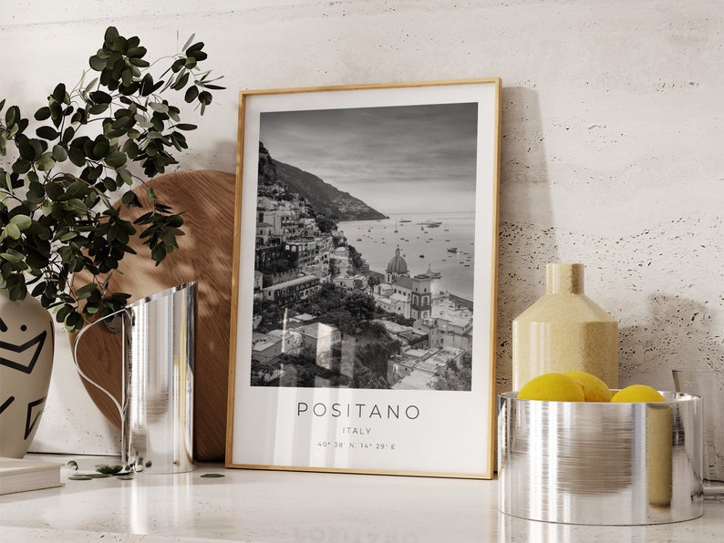 May include: Black and white photograph of Positano, Italy, framed in a light wood frame. The photo shows a coastal town with buildings on a hillside overlooking the sea. The text "Positano, Italy" is printed below the photo.