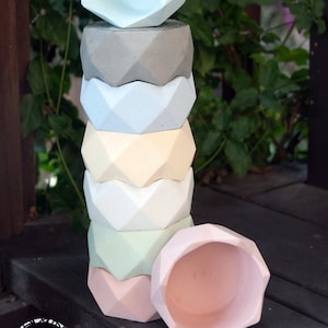 May include: A stack of seven geometric concrete planters in various pastel colors, including light blue, gray, cream, light green, and pink. The planters are stacked on a wooden surface.