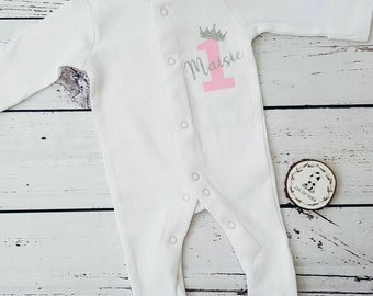 May include: White baby onesie with a pink number one and a silver crown. The name Maisie is written in silver glitter on the onesie.