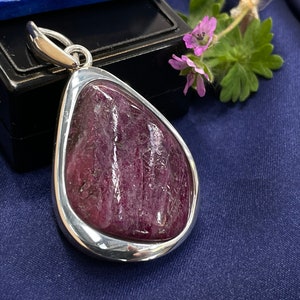 Puede incluir: Colgante de plata con una piedra preciosa roja oscura en forma de lágrima. La piedra está engastada en un bisel plateado. El fondo es de tela azul oscuro, con una ramita de flores moradas. Joya.