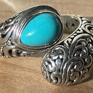 Peut inclure: Une bague en argent ornée d'une pierre ovale turquoise vif. La bague présente un motif tourbillonnant complexe autour de la pierre et sur le corps. La bague est présentée sur une surface brun clair.