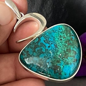 Peut inclure: Un pendentif en argent avec une grosse pierre triangulaire polie. La pierre présente un mélange de couleurs turquoise et vert foncé. Le pendentif a une bélière incurvée pour une chaîne de collier. Une fleur violette est en arrière-plan.