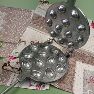 Könnte beinhalten: Ein Waffeleisen aus silbernem Metall mit einem langen Griff. Das Eisen hat zwei runde Abschnitte, jeder mit 12 Formen in Walnussform. Das Eisen ist geöffnet und zeigt das Innere der Formen.