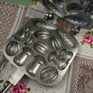 May include: A vintage metal mold for making candy or chocolate. The mold has a variety of shapes, including mushrooms, nuts, and other designs. The mold is made of silver-colored metal and has a handle.