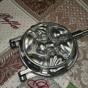 Op de afbeelding: Een zilverkleurige, vintage koekjesvorm met verschillende vormen, waaronder harten en schelpen. De vorm heeft een lange handgreep en is ontworpen voor het maken van decoratieve koekjes. De vorm is van metaal en wordt waarschijnlijk gebruikt om te bakken.