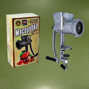 May include: A silver manual meat grinder with a black handle. The grinder is attached to a table with a clamp. The box for the grinder is in the background and says "Мясорубка" in Russian.
