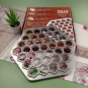 May include: A silver metal mold with 42 round indentations arranged in a hexagonal pattern. The mold is used for making dumplings. The mold is on a table with a floral pattern tablecloth.