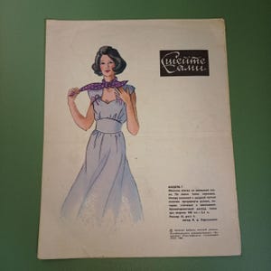 Op de afbeelding: Een vintage naaipatroon met een vrouw die een lichtblauwe jurk draagt met een paarse stippen sjaal. Het patroon is getiteld "Sheite Sami" en bevat instructies voor het maken van de jurk.
