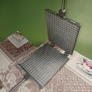 May include: A vintage, open waffle iron made of silver-colored metal. The iron features a grid pattern on both cooking surfaces and a long handle. The waffle iron is resting on a floral patterned tablecloth.