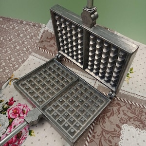 Op de afbeelding: Een zilveren wafeltang met een scharnierend deksel en een lange handgreep. De wafeltang heeft twee secties, elk met een rasterpatroon van vierkanten.