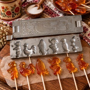 May include: A metal candy mold featuring six distinct animal shapes including a bear, rooster, fish, elephant, penguin, and rabbit. The mold is positioned above six matching amber-colored lollipops on sticks, each reflecting the corresponding animal design.