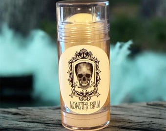 Puede incluir: Un recipiente cilíndrico transparente de Monster Balm con una etiqueta de calavera. La etiqueta ovalada tiene un marco ornamental en blanco y negro alrededor de una calavera. El texto "MONSTER BALM" está debajo. El texto "NEW 1/2oz size" está en la parte inferior.