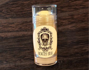 Puede incluir: Un recipiente cilíndrico transparente de Monster Balm con un bálsamo de color crema en su interior. La etiqueta presenta una calavera en un marco decorativo y las palabras "MONSTER BALM". El recipiente está sobre una superficie de madera oscura.