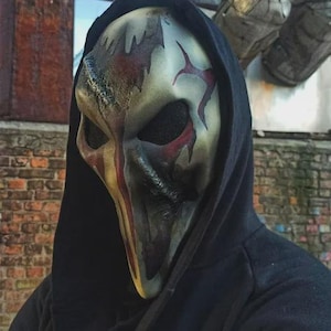 May include: A detailed, full-face mask with a distressed, aged appearance. The mask features a pale base color with red and black accents, including streaks and stylized markings. It is worn with a black hooded garment, creating a mysterious look.