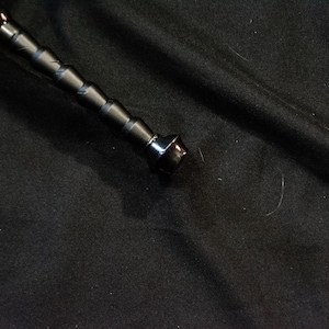 Puede incluir: Una espada negra con una empu&ntilde;adura texturizada y una hoja plateada. La empu&ntilde;adura de la espada tiene una serie de patrones en forma de diamante en relieve. La hoja es curva y tiene una punta afilada. La espada est&aacute; sobre una superficie negra.