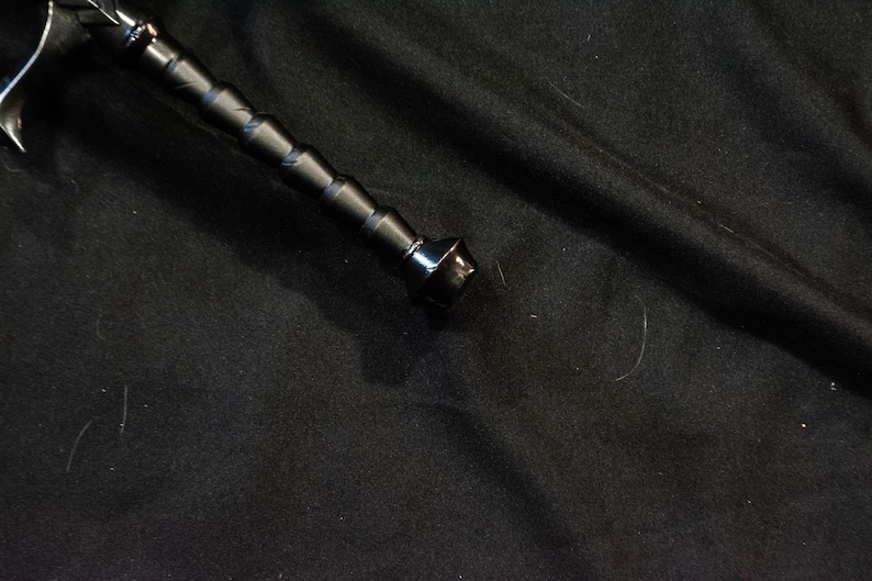 Puede incluir: Una espada negra con una empu&ntilde;adura texturizada y una hoja plateada. La empu&ntilde;adura de la espada tiene una serie de patrones en forma de diamante en relieve. La hoja es curva y tiene una punta afilada. La espada est&aacute; sobre una superficie negra.