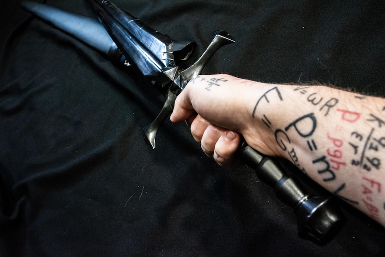 Puede incluir: Una espada negra con una hoja oscura y angular y un mango negro. La espada se sostiene en una mano con ecuaciones matem&aacute;ticas tatuadas en el brazo. El dise&ntilde;o de la espada sugiere un tema de fantas&iacute;a o hist&oacute;rico.