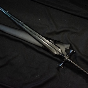 Puede incluir: Una espada negra con una hoja larga y delgada y una empu&ntilde;adura detallada. La espada est&aacute; sobre una superficie negra, con una vaina parcialmente visible. El dise&ntilde;o presenta una guarda y un mango texturizado.