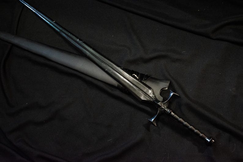 Puede incluir: Una espada negra con una hoja larga y delgada y una empu&ntilde;adura detallada. La espada est&aacute; sobre una superficie negra, con una vaina parcialmente visible. El dise&ntilde;o presenta una guarda y un mango texturizado.