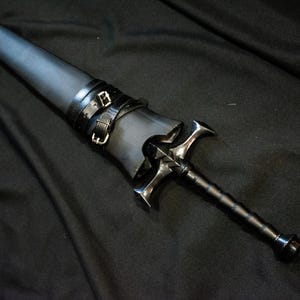 Puede incluir: Una espada negra con una empu&ntilde;adura plateada en forma de cruz y un mango negro. La espada est&aacute; enfundada en una vaina de cuero negro asegurada con dos correas de cuero negro y hebillas plateadas. El fondo es una tela oscura.