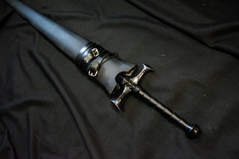 Puede incluir: Una espada negra con una empu&ntilde;adura plateada en forma de cruz y un mango negro. La espada est&aacute; enfundada en una vaina de cuero negro asegurada con dos correas de cuero negro y hebillas plateadas. El fondo es una tela oscura.
