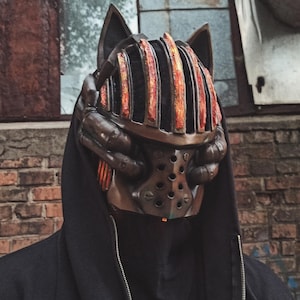 May include: A detailed, steampunk-inspired mask with cat-like ears. The mask features a metallic finish with orange and brown accents, a cage-like structure over the face, and multiple rivets. The person is wearing a black hooded sweatshirt.
