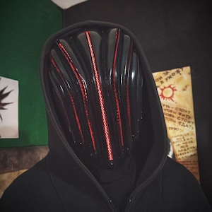 May include: A black mask with vertical red detailing, worn with a black hooded top. The mask features a futuristic design, with parallel red lines. The overall aesthetic is dark and mysterious.