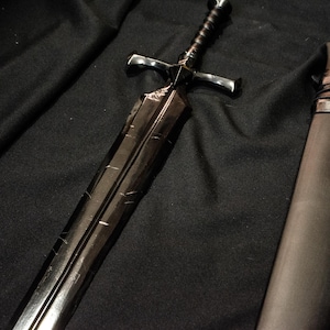 Puede incluir: Una espada negra con una empu&ntilde;adura en forma de cruz y un mango texturizado descansa sobre una superficie oscura. La hoja tiene l&iacute;neas grabadas y un acabado oscuro. Una vaina negra con correas est&aacute; al lado de la espada.