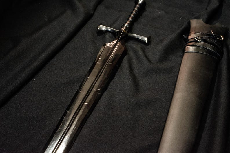 Puede incluir: Una espada negra con una empu&ntilde;adura en forma de cruz y un mango texturizado descansa sobre una superficie oscura. La hoja tiene l&iacute;neas grabadas y un acabado oscuro. Una vaina negra con correas est&aacute; al lado de la espada.