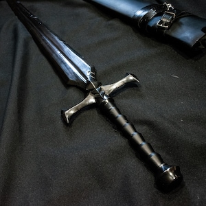 Puede incluir: Una espada negra y plateada con una guarda y un mango texturizado. La hoja es oscura y reflectante. Una vaina negra con hebilla est&aacute; en el fondo. La espada est&aacute; sobre una superficie negra.