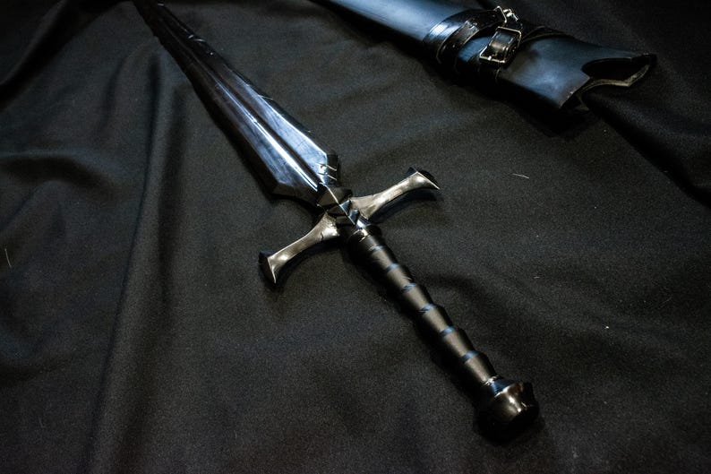 Puede incluir: Una espada negra y plateada con una guarda y un mango texturizado. La hoja es oscura y reflectante. Una vaina negra con hebilla est&aacute; en el fondo. La espada est&aacute; sobre una superficie negra.