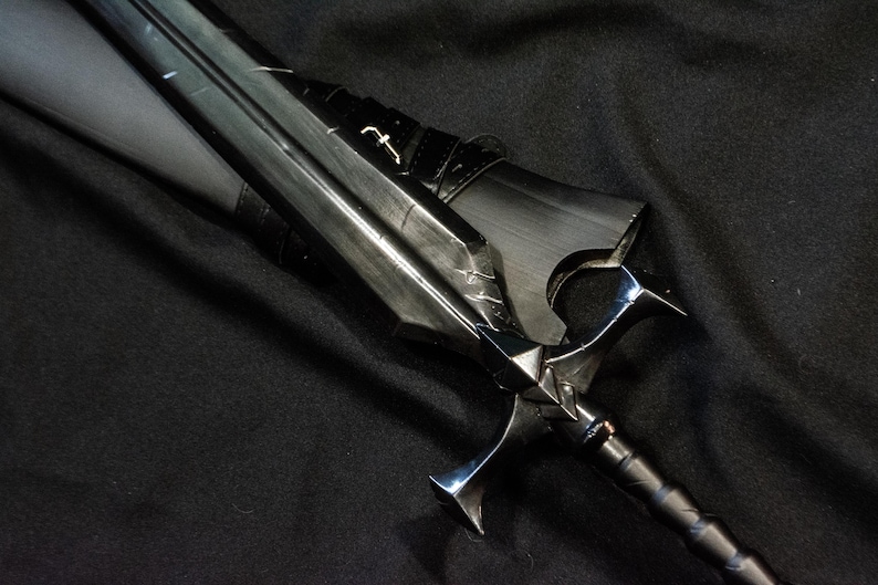 Puede incluir: Una espada negra con una guarda plateada y una hoja detallada. La espada tiene un mango oscuro y est&aacute; sobre un fondo negro. La hoja tiene dise&ntilde;os grabados y un peque&ntilde;o emblema.