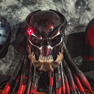 May include: A detailed Predator mask with a metallic finish, featuring a red targeting laser and intricate details. The mask has a skeletal jawline and dreadlock-like tendrils with red tips, showcasing a sci-fi theme.