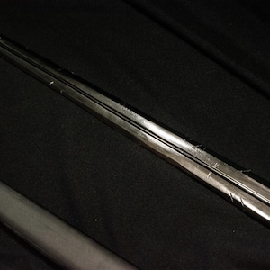 Puede incluir: Un objeto met&aacute;lico largo, similar a una espada, con una cresta central y una superficie oscura y reflectante. El objeto est&aacute; colocado en diagonal sobre un fondo oscuro, lo que sugiere un arma o un objeto decorativo.