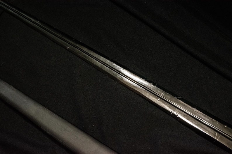 Puede incluir: Un objeto met&aacute;lico largo, similar a una espada, con una cresta central y una superficie oscura y reflectante. El objeto est&aacute; colocado en diagonal sobre un fondo oscuro, lo que sugiere un arma o un objeto decorativo.