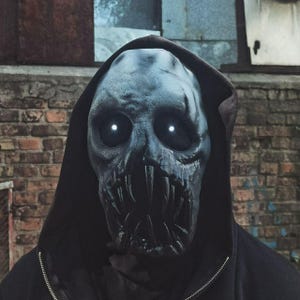 Op de afbeelding: Een gedetailleerd, grijs monster masker met gloeiende witte ogen en scherpe tanden. Het masker wordt gedragen met een zwarte hoodie, wat een dreigende uitstraling creëert. De achtergrond toont een bakstenen muur en een glimp van een raam.
