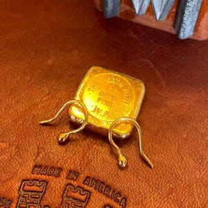 May include: Two gold-colored earring hooks on a brown leather surface. The hooks are made of metal and have a simple design. A small gold-colored square with the text "10K 585 FINE .W.P." is visible in the background.