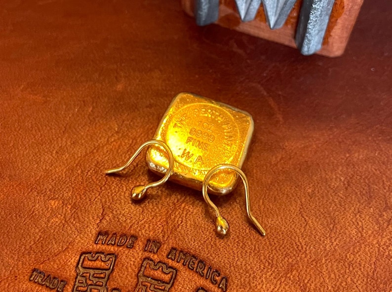 May include: Two gold-colored earring hooks on a brown leather surface. The hooks are made of metal and have a simple design. A small gold-colored square with the text "10K 585 FINE .W.P." is visible in the background.