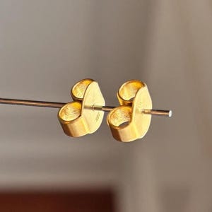 Könnte beinhalten: Nahaufnahme von zwei goldfarbenen Ohrringverschlüssen. Die Ohrringverschlüsse sind schmetterlingsförmig und haben eine glänzende Oberfläche. Die Ohrringe befinden sich auf einem dünnen Metallstab, und der Hintergrund ist eine verschwommene neutrale Farbe.